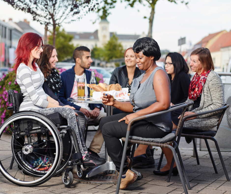 An einem Tisch sitzen eine Person im Rollstuhl und andere mit verschiedenen Migrationshintergründen und unterhalten sich lachend