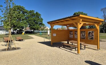 Chillplatz Schönkirchen-Reyersdorf (C) LEADER Region Weinviertel Ost Überdachter Holzpavillon mit Sitzgelegenheiten auf einem neu gestalteten Aufenthaltsplatz. Daneben befinden sich Liegebänke, junge Bäume und Wege aus hellem Schotter. Im Hintergrund sind Grünflächen und Spielgeräte sichtbar.