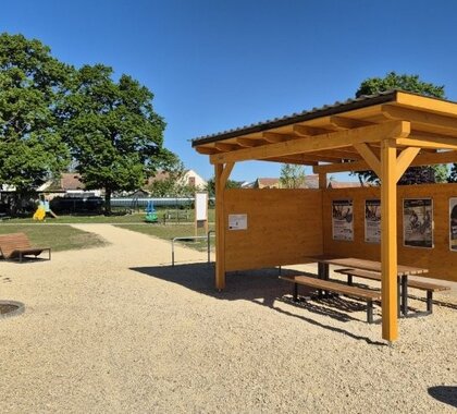 Überdachter Holzpavillon mit Sitzgelegenheiten auf einem neu gestalteten Aufenthaltsplatz. Daneben befinden sich Liegebänke, junge Bäume und Wege aus hellem Schotter. Im Hintergrund sind Grünflächen und Spielgeräte sichtbar.