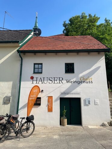 Das Presshaus der Familie Hauser von außen, Blick von der Poysdorfer Kellergstetten. Ein zweistöckiges Presshaus mit einer offenen Kellertüre im Erdgeschoss, zwei kleinen Fenstern im Obergeschoss. Vor dem Presshaus steht ein Weinviertel Beachflag sowie 2 Fahrräder.