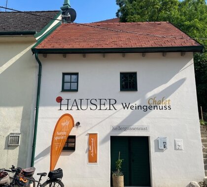 Das Presshaus der Familie Hauser von außen, Blick von der Poysdorfer Kellergstetten. Ein zweistöckiges Presshaus mit einer offenen Kellertüre im Erdgeschoss, zwei kleinen Fenstern im Obergeschoss. Vor dem Presshaus steht ein Weinviertel Beachflag sowie 2 Fahrräder.
