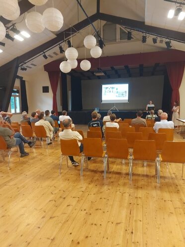 Bürgerinfoveranstaltung zum Teilbebauungsplan in Falkenstein: Mehrere Personen sitzen auf Stühlen im Veranstaltungssaal mit Holzfußboden und schauen in Richtung Bühne. Auf der Bühne steht eine Person neben einer Leinwand und hält eine Präsentation. Über der Bühne hängen runde Lampions.