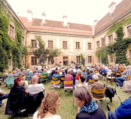 Besucherinnen und Besucher sitzen bei einer Kulturveranstaltung im begrünten Innenhof des historischen Banabiten Klosters in Mistelbach und verfolgen ein Konzert vor der Kulisse der alten Klostergebäude. Der Innenhof ist voll besetzt, es sind viele Menschen zu sehen.
