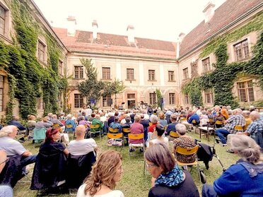 Besucherinnen und Besucher sitzen bei einer Kulturveranstaltung im begrünten Innenhof des historischen Banabiten Klosters in Mistelbach und verfolgen ein Konzert vor der Kulisse der alten Klostergebäude. Der Innenhof ist voll besetzt, es sind viele Menschen zu sehen.