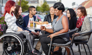 An einem Tisch sitzen eine Person im Rollstuhl und andere mit verschiedenen Migrationshintergründen und unterhalten sich lachend