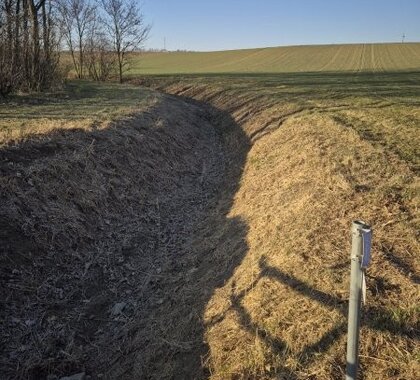 Wasserführender Graben der Gemeinde Gaubitsch in dem die Staubauwerke errichtet werden, rechts ist ein Acker zu sehen, links eine Verbuschung, mittig ein Graben ohne Wasser der in der Ferne nach links schwenkt