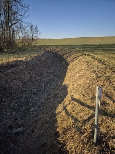 Wasserführender Graben der Gemeinde Gaubitsch in dem die Staubauwerke errichtet werden, rechts ist ein Acker zu sehen, links eine Verbuschung, mittig ein Graben ohne Wasser der in der Ferne nach links schwenkt