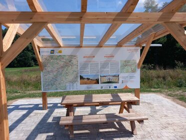 Der Weinviertel Rastplatz in der Gemeinde Wilfersdorf, unter einer Pergula befindet sich eine Tisch-Bank-Kombination mit dahinterliegender Informationstafel über den Radweg.