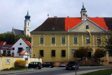 Historisches Klostergebäude in Mistelbach mit barocker Fassade und rotem Dach; im Hintergrund sind Kirchturm der Stadtpfarrkirche und weitere Gebäude des Klosterensembles zu sehen.