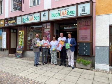 Am Bild zu sehen die Vertreter der Projektträger Städte vor einem Buchladen mit dem Werbesujet Plakat zur Cities-App