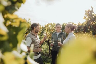 Ein junger Winzer steht mit zwei Frauen im Weingarten. Alles drei Personen halten ein Weinglas in der Hand, der Winzer zusätzlich eine Weinfalsche.