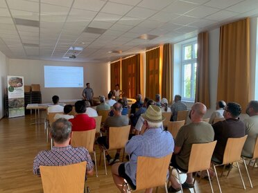 Bei der Infoveranstaltung zur Kellergassen Schutzzone Wildendürnbach Präsentiert der Raumplaner , davor sitzen rund 20 Personen auf Stühlen und hören dem Vortrag zu.