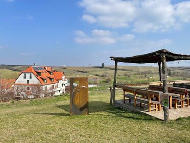 Ein beschatteter Rastplatz mit Tischen und Bänken, daneben eine Landmark mit dem Neustifter-Logo. Im Hintergrund ist das Hotel und die Weinviertler Landschaft zu sehen.