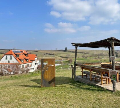 Ein beschatteter Rastplatz mit Tischen und Bänken, daneben eine Landmark mit dem Neustifter-Logo. Im Hintergrund ist das Hotel und die Weinviertler Landschaft zu sehen.