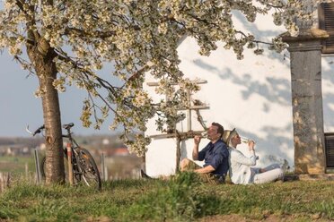 Ein junges Pärchen sitzt vor einem Weinkeller unter einem blühenden Baum und genießt ein Achterl Wein.