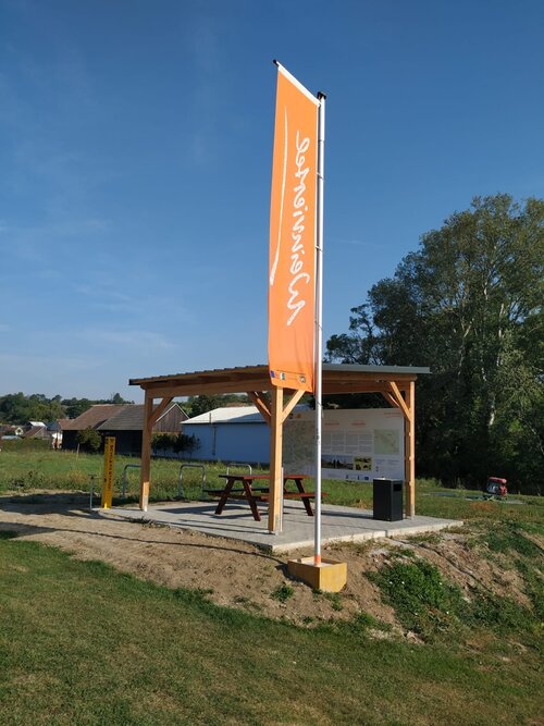 Ein Weinviertel Radrastplatz in der Landschaft mit einer Holzpergola und einer orangenen Weinviertel Fahne davor.