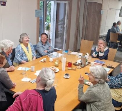 Eine Gruppe von rund zehn älteren Frauen sitzt an einem großen Tisch in einem Veranstaltungsraum und tauscht sich bei Kaffee und Gebäck aus. Die Teilnehmerinnen sind in Gespräch vertieft und verbringen gemeinsam einen geselligen Nachmittag. Die Atmosphäre wirkt offen, freundlich und gemeinschaftlich.