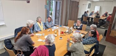 Caring Region Wolkersdorf (C) Caring Region Wolkersdorf Eine Gruppe von rund zehn älteren Frauen sitzt an einem großen Tisch in einem Veranstaltungsraum und tauscht sich bei Kaffee und Gebäck aus. Die Teilnehmerinnen sind in Gespräch vertieft und verbringen gemeinsam einen geselligen Nachmittag. Die Atmosphäre wirkt offen, freundlich und gemeinschaftlich.