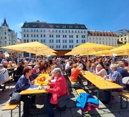 In Wien am Hof fand 2023 das Weinviertel Fest statt. Zu sehen sind viele Personen die an Biertischen sitze, unter orangen Liegestühlen in der Kulisse der Gebäude vom Wiener Hof.