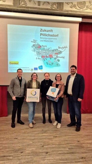 Präsentation Masterplan Pillichsdorf Fünf Personen präsentieren bei einer Veranstaltung den Beteiligungsprozess und Masterplan „Zukunft Pillichsdorf“. Im Hintergrund ist eine Präsentationsfolie mit einer Illustration des Ortszentrums zu sehen.