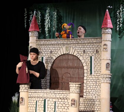 Zwei Puppenspieler führen mit einer bunten Figur ein Theaterstück vor einer großen, mit Türmen verzierten Burgkulisse auf.