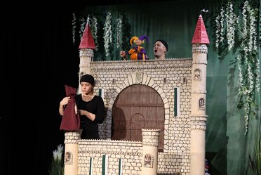 Puppentheater_(c)Markus Mathes Zwei Puppenspieler führen mit einer bunten Figur ein Theaterstück vor einer großen, mit Türmen verzierten Burgkulisse auf.