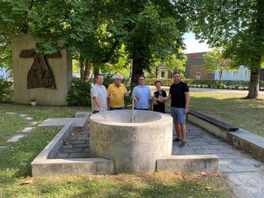 Eine Gruppe von 5 Personen (4 Bürgermeister:innen und Alexander Wimmer als Kleinregionsmanager) steht vor einem betonierte Brunnen aus dem Wasser rinnt. Die Personen halten Trinkgläser sowie einen Krug mit Wasser in der Hand.
