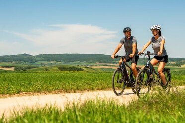 Eine Radfahrerin und ein Radfahrer fahren auf einem Radweg zwischen Feldern, im Hintergrund ist der Buschberg zu sehen.