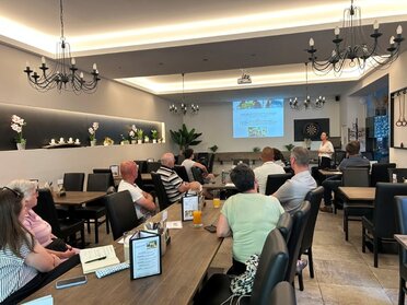 Präsentation Sozialraumanalyse Leiser Berge, C: REV Leiser Berge Eine Dame präsentiert die Ergebnisse der Sozialraumanalyse, an der Leinwand im Hintergrund ist die Studie zu sehen. Im Vordergrund sitzen Personen an Tischen mit dem Blick auf die Leinwand gerichtet.