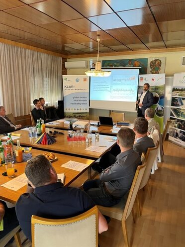 Gemeindevertreter:innen sitzen um einen Arbeitstisch. Im vorderen des Raumes steht eine Präsentationswand auf der Ergebnisse der Befragung gezeigt werden. Ein Person steht neben der Präsentationswand und informiert die anwesenden Personen.