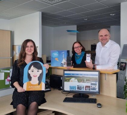 Bianca Christenheit (hält den Dummy für den Chatbot in der Hand), Christine Filipp und Christian Frank welche den Chatbot am Handy offen haben. Im Vordergrund steht ein Bildschirm auf welchem die Webseite der Gemeinde Herrnbaumgarten aufgerufen ist und eine Frage im Chatbot gestellt wird.