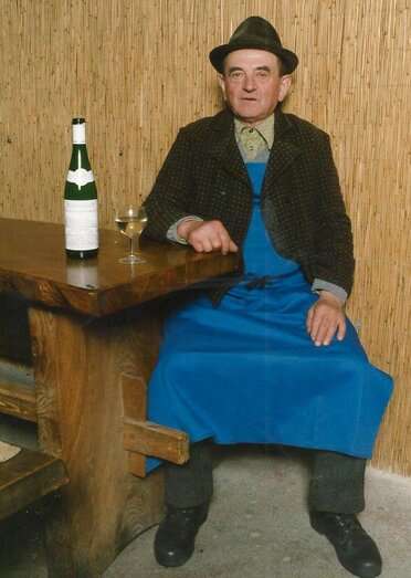 Ein äterer Kellermann sitzt in einem Presshaus an einem Holztisch. Er trägt typisches Weinviertler Gewans mit einem Firta (blaue Arbeitsschürze), einen Kalmuk (traditionelle Jacke) sowie einen Hut. Auf dem Tisch steht eine Weinflasche und ein Achtel mit Weißwein.