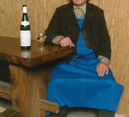 Ein äterer Kellermann sitzt in einem Presshaus an einem Holztisch. Er trägt typisches Weinviertler Gewans mit einem Firta (blaue Arbeitsschürze), einen Kalmuk (traditionelle Jacke) sowie einen Hut. Auf dem Tisch steht eine Weinflasche und ein Achtel mit Weißwein.