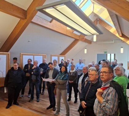 Diskussionsformat in Pillichsdorf zum neuen Teilbebauungsplan. Viele Bewohner:innen stehen im Gemeindesaal mit dem Blick richtung Präsentationsfläche. Am Rand stehen Pinnwände auf denen Pläne aufgehängt sind.