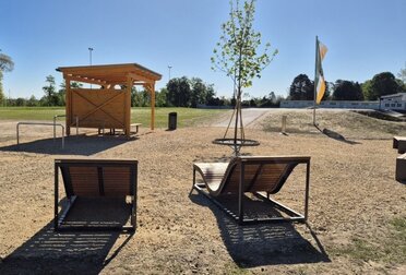 Chillplatz Schönkirchen-Reyersdorf (C) LEADER Region Weinviertel Ost Neu gestalteter Aufenthaltsbereich im Freien mit überdachtem Holzsitzplatz, zwei modernen Liegebänken, einem jungen Baum und befestigten Wegen. Die Anlage liegt auf einer offenen Grünfläche und bietet Möglichkeiten zum Ausruhen und Verweilen.