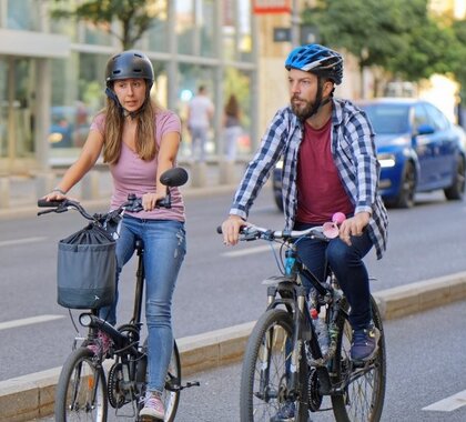 Auf dem Symbolbild, dass das Alltagsradfahren im Weinviertel darstellen sollen, ist eine Frau und ein Mann zu sehen, welche mit einem Fahrrad in einer Stadt fahren. Beide tragen einen Helm und sind in Jeans und T-Shirt unterwegs. Sie fahren auf einer Radspur, im Hintergrund ist ein Auto auf der danebenliegenden Straße zu sehen.